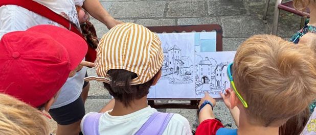 Eine Gruppe von Kindern und eine erwachsene Frau sind um eine Zeichnung auf einem Brett versammelt. Sie tragen alle Mützen und Rucksäcke. Die Frau trägt eine Sonnenbrille und eine rote Umhängetasche.