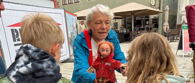 Eine ältere Frau zeigt zwei Kindern in einer Stadt einen Puppen in einem roten Regenmantel. Die Kinder tragen Jacken. Im Hintergrund Gebäude und ein Regenschirm.
