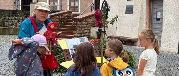 Eine Frau und mehrere Kinder stehen vor einem Gebäude mit dem Schriftzug Stille Nacht Museum Hallen. Die Frau hält ein Gemälde und eine Puppe. Die Kinder tragen Regenmäntel und Rucksäcke.
