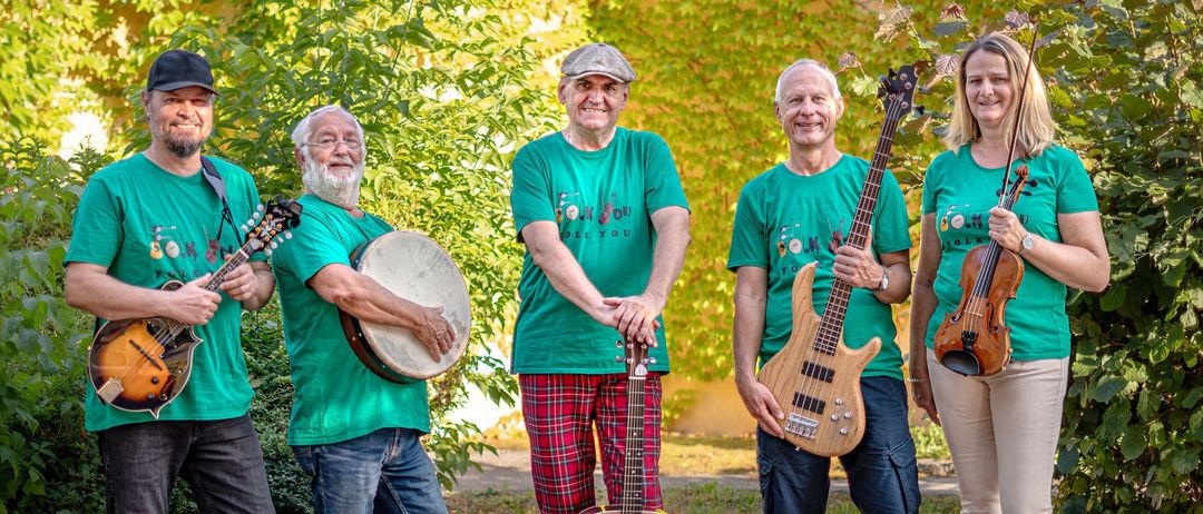 Drei Männer stehen lächelnd im Freien. Der linke Mann hält eine Trommel, der mittlere eine Gitarre und der rechte einen Bassgitarre. Sie stehen vor einem Busch.