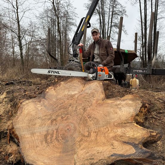 Ein Mann in einer braunen Jacke und einem Hut hält eine Kettensäge vor einem gefällten Baum im Wald.