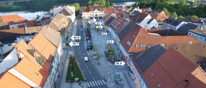 Ein Luftbild einer Stadt mit nummerierten Straßen. Gebäude säumen beide Seiten, einige mit braunen Dächern. Eine Straße verläuft mit geparkten Autos und einem Fußgängerüberweg. Bäume und Grünflächen sind vorhanden.