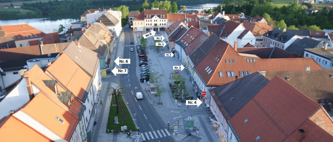 Ein Luftbild einer Stadt mit nummerierten Straßen. Gebäude säumen beide Seiten, einige mit braunen Dächern. Eine Straße verläuft mit geparkten Autos und einem Fußgängerüberweg. Bäume und Grünflächen sind vorhanden.