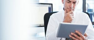 Ein konzentrierter Mann sitzt an einem Schreibtisch und schaut auf ein Tablet, während er formelle Kleidung trägt.