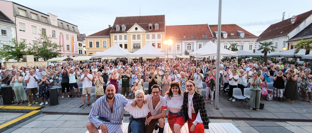 Fünf Personen posieren auf einer Bühne vor einem großen Publikum in einem Freiluft-Veranstaltungsbereich mit Zelten, Gebäuden und Bäumen im Hintergrund.