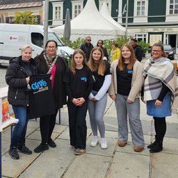 Eine Gruppe von Frauen und Mädchen steht vor einem Gebäude und lächelt für die Kamera. Eine hält ein schwarzes T-Shirt mit 'Give' drauf. Dahinter ist ein Lieferwagen geparkt und ein Zelt aufgebaut.