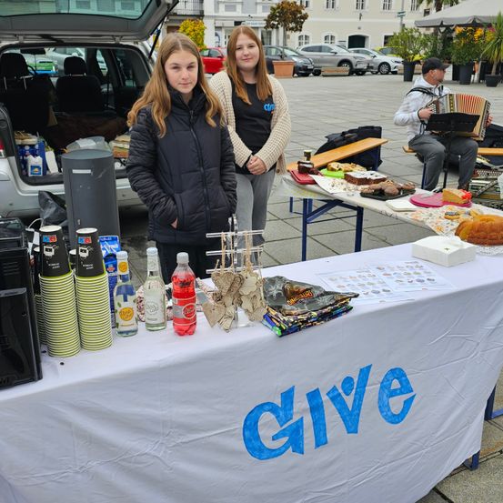 Zwei Mädchen stehen hinter einem Tisch mit dem Wort 'geben' darauf und bieten verschiedene Artikel zum Verkauf an. Ein Mann spielt Akkordeon in der Nähe, und andere Autos sind um sie herum geparkt.
