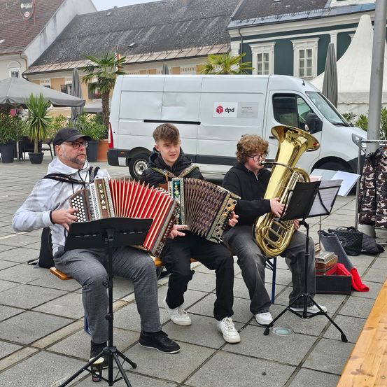 Drei Musiker spielen auf einem Stadtplatz, einer sitzt auf einem Stuhl und spielt Akkordeon, ein anderer sitzt auf einem Hocker und spielt Tuba, und ein Dritter sitzt auf einer Bank und spielt Tuba.