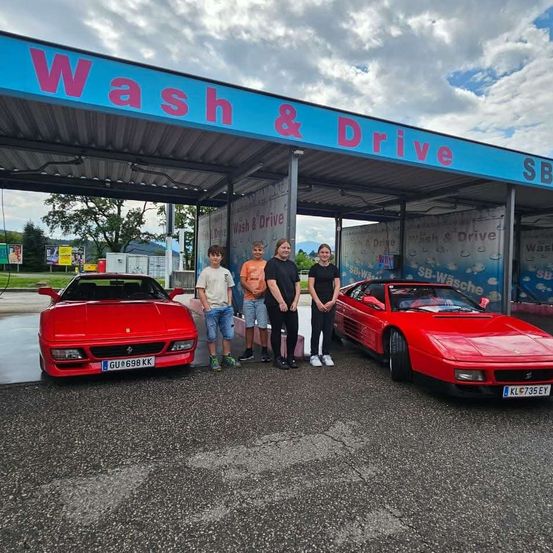 Fünf Personen stehen unter einem 'Wash & Drive'-Schild, mit zwei roten Ferraris vor einer Autowaschanlage geparkt.