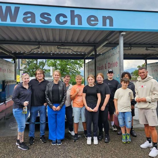 Eine Gruppe von Menschen steht vor einer Autowaschanlage und lächelt für ein Foto. Das Schild über ihnen lautet 'Waschen'. Bäume und Berge sind im Hintergrund zu sehen.