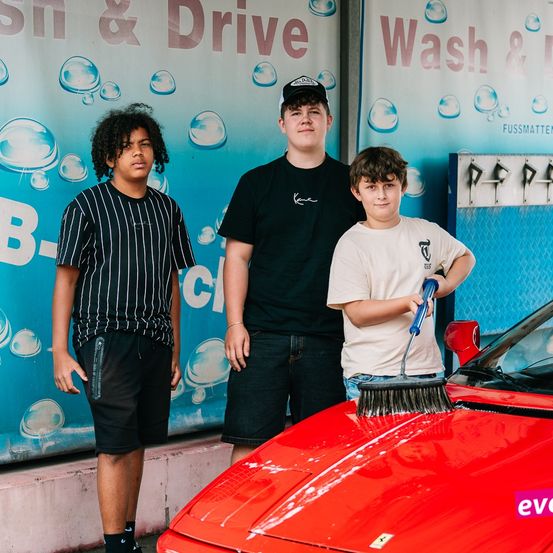Drei Jungen stehen vor einem roten Auto, einer mit einer Bürste, ein anderer mit einem Tuch. Hinter ihnen befindet sich ein Banner mit Wash & Drive.