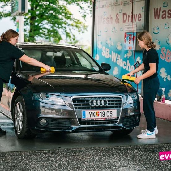 Zwei Frauen reinigen einen schwarzen Audi an einer Autowaschanlage. Eine reinigt die Windschutzscheibe, die andere den Seitenspiegel. Ein Schild der Autowaschanlage befindet sich hinter ihnen.