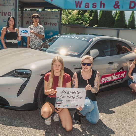 Zwei Frauen und ein Mädchen posieren vor einem weißen Auto mit einem Schild, das Volkswagen Car Wash Day steht. Sie halten ein weißes Schild, das Give sagt. Dahinter befindet sich eine Autowaschanlage mit einem Banner, das Wash & Drive sagt.