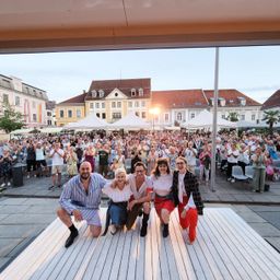 Fünf Personen stehen auf einer hölzernen Bühne in einem Stadtplatz mit einem Publikum. Die Leute klatschen und lächeln. Im Hintergrund sind Gebäude zu sehen.