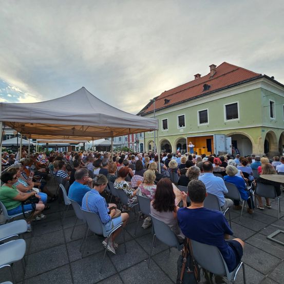 Eine Veranstaltung im Freien mit vielen Menschen, die auf Stühlen sitzen. Eine Person steht auf der Bühne und hält eine Rede. In der linken Ecke steht ein großes Zelt. Der Himmel ist bewölkt.
