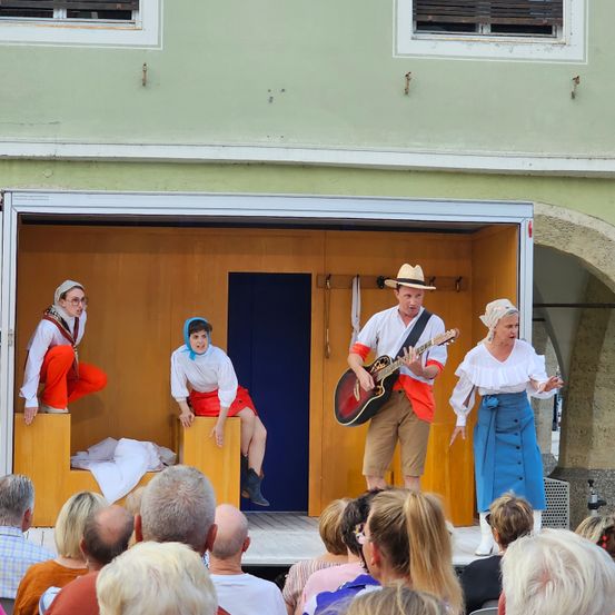 Eine Gruppe von Darstellern auf einer Bühne mit einem Mann, der eine Gitarre spielt, und zwei Frauen, die auf hölzernen Bänken sitzen, vor einem Publikum.