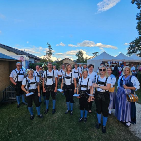 Eine Gruppe von Musikern in traditioneller Kleidung posiert für ein Foto auf einem Rasenfeld. Hinter ihnen sind Zelte und Gebäude zu sehen.