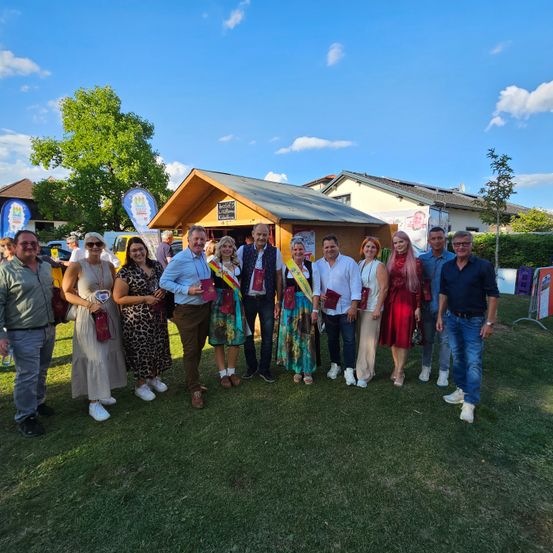 Eine Gruppe von Menschen steht im Freien und lächelt für ein Foto. Sie tragen Freizeitkleidung, einige mit Schärpen. Ein kleines Gebäude und Bäume sind im Hintergrund, unter einem klaren blauen Himmel.