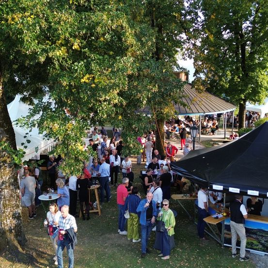 Eine Freiluftveranstaltung mit vielen Menschen unter Zelten, Tischen und Bäumen. Einige unterhalten sich, andere sitzen, und ein Mann hält ein Mikrofon.