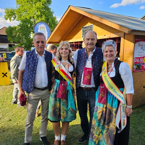 Drei Erwachsene und eine junge Frau stehen vor einer Holzbude mit einem Schild, möglicherweise auf einem Festival. Sie tragen alle traditionelle Kleidung und lächeln für die Kamera. Hinter ihnen stehen weitere Personen und ein gelber Mülleimer.