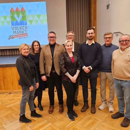 Eine Gruppe von Menschen steht drinnen und lächelt für ein Foto. Sie sind lässig gekleidet. Eine große Leinwand hinter ihnen zeigt den Text 'Volker Markt 20 Jahre.'