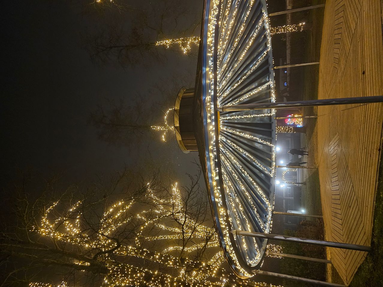 Ein Karussell ist nachts beleuchtet, mit vielen kleinen Lichtern an den Bäumen und Gebäuden. Das Karussell ist mit bunten Lichtern dekoriert und befindet sich scheinbar in der Mitte eines Parks.