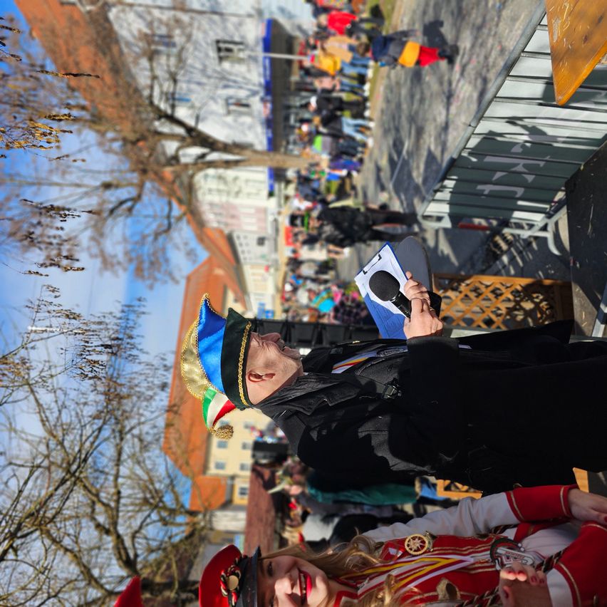 Ein Mann in traditioneller Kleidung hält ein Mikrofon und spricht möglicherweise zu einer Menschenmenge in einer Freiluftveranstaltung mit Gebäuden und Bäumen im Hintergrund.