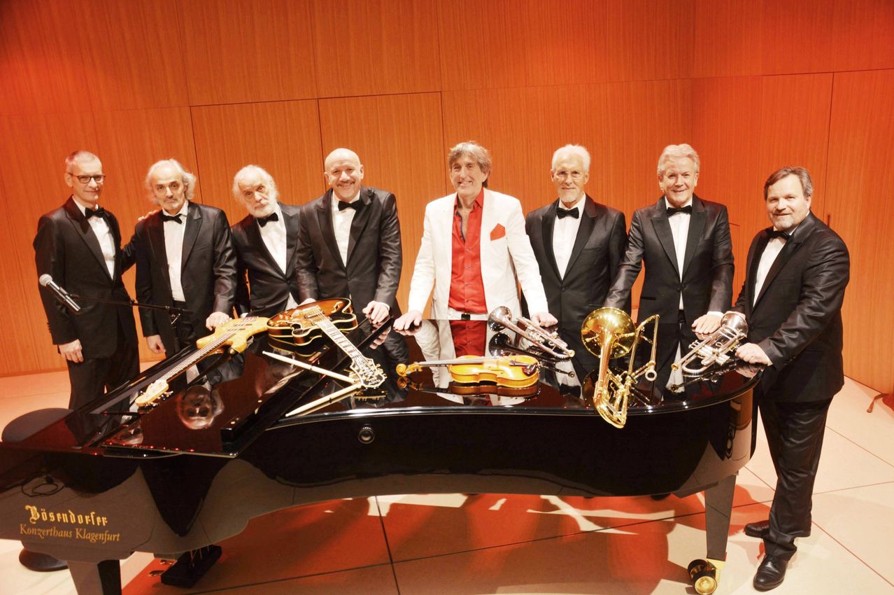 Sieben Männer stehen hinter einem Klavier, alle in Anzügen und Fliegen. Jeder hält ein Musikinstrument. Der Mann in der Mitte trägt ein rotes Hemd.