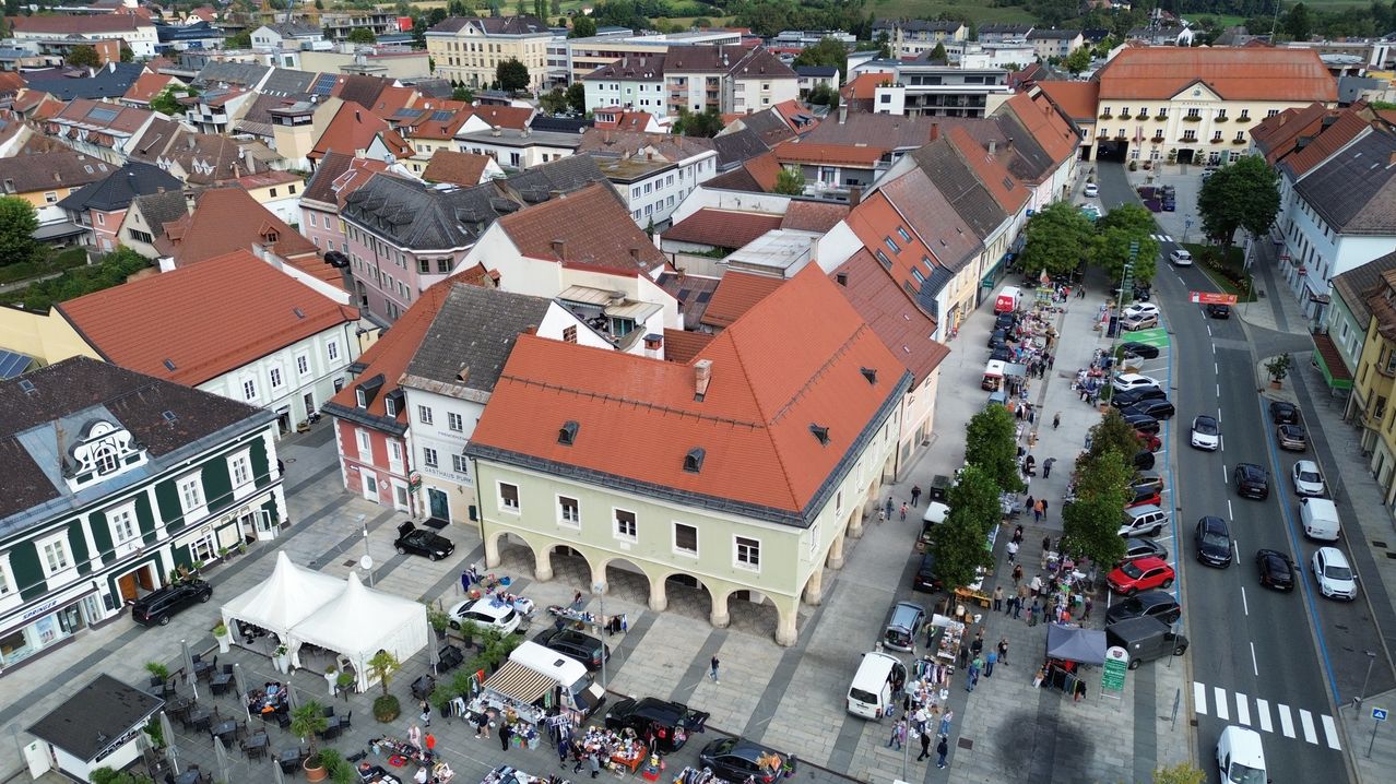 Ein Luftbild eines geschäftigen Stadtplatzes. Viele Menschen gehen, und mehrere Autos sind geparkt. Es gibt Stände, die Gegenstände verkaufen, und ein großes Gebäude mit rotem Dach dominiert die Szene. Bäume und Grünflächen sind rund um den Platz verteilt.