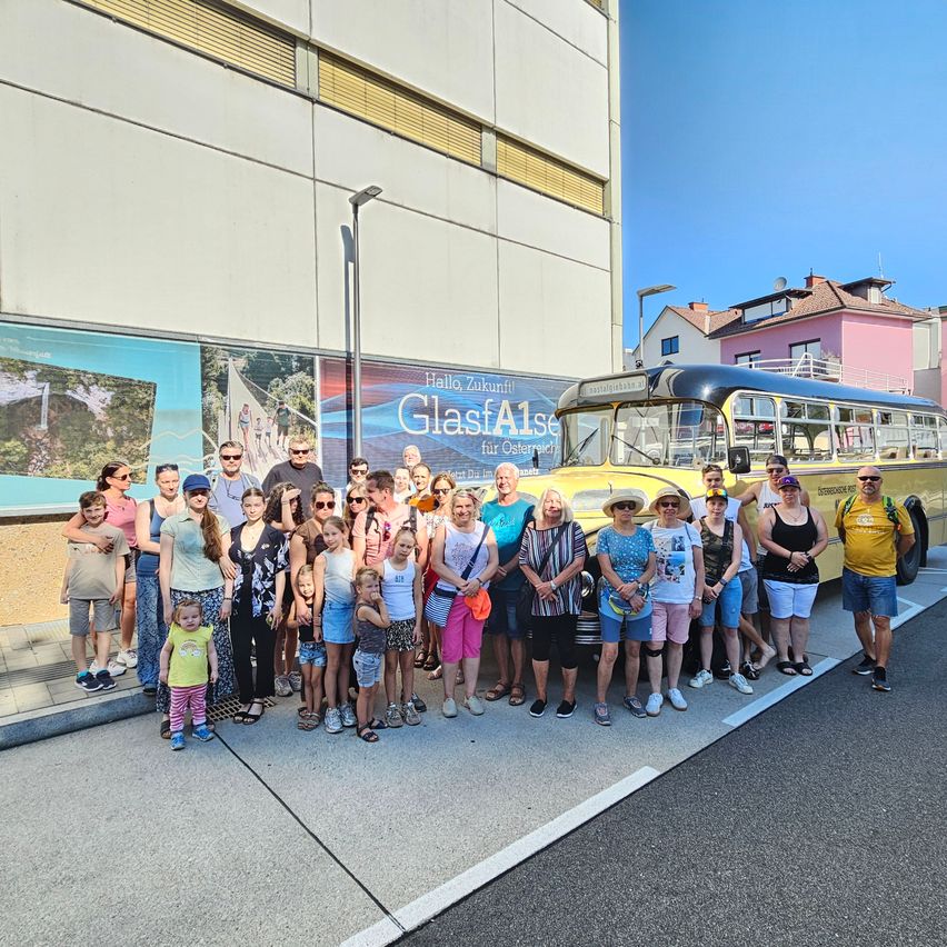 Eine Gruppe von Menschen, einschließlich Erwachsener und Kindern, posiert für ein Foto vor einem gelben Bus mit der Aufschrift 'GlasAls' an der Seite. Sie stehen auf dem Bürgersteig, mit einem Gebäude und einer Straßenlaterne im Hintergrund.