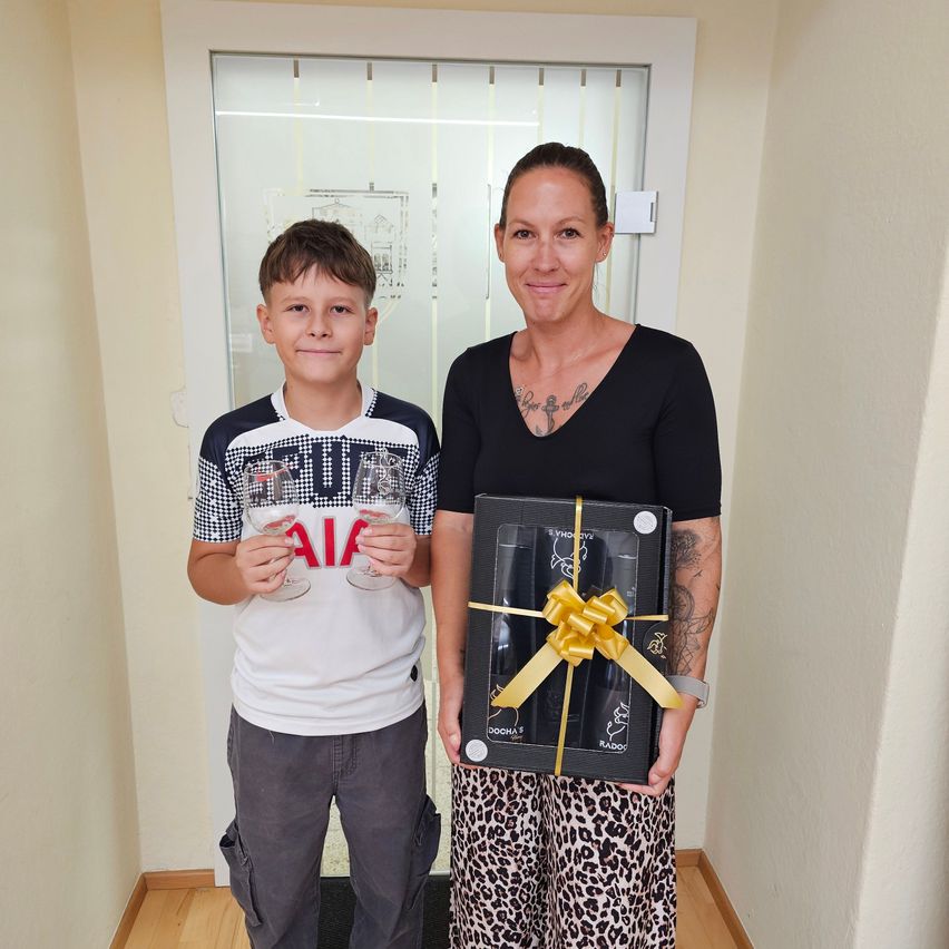 Eine Frau und ein Junge stehen vor einer Glastür und lächeln. Die Frau hält eine Geschenkbox mit einem Band und der Junge hält zwei Weingläser.