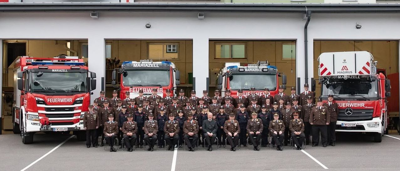 Eine Gruppe Feuerwehrleute in Uniform posiert für ein Foto vor ihrer Feuerwache, mit mehreren Feuerwehrautos im Vordergrund geparkt.