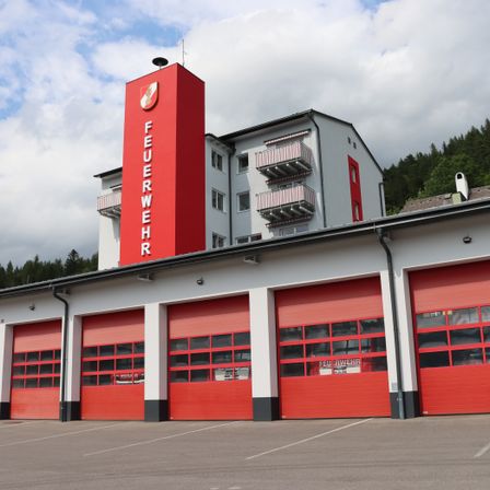 Ein modernes Feuerwehrhaus mit einem roten Turm und großen roten Garagentoren, vor dem Hintergrund von bewölktem Himmel und bewaldeten Hügeln.