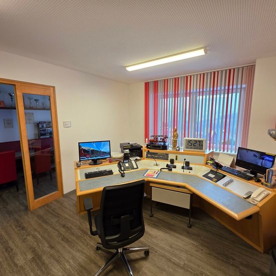 Ein Büro mit Schreibtisch, Computerbildschirmen und Bürogeräten. Der Raum hat Holzböden und weiße Wände.
