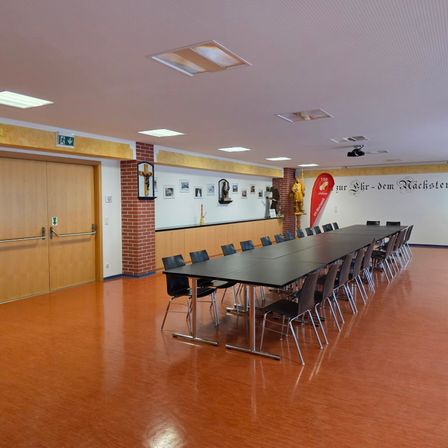Ein leerer Konferenzraum mit langen schwarzen Tischen und Stühlen, Holztüren und einer weißen Wand mit Bildern und einem roten Banner.