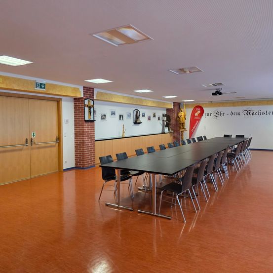 Ein leerer Konferenzraum mit langen schwarzen Tischen und Stühlen, Holztüren und einer weißen Wand mit Bildern und einem roten Banner.