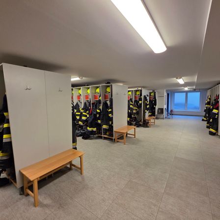 Ein leerer Feuerwehrumkleideraum mit Reihen von Schließfächern, Helmen und Uniformen. Holzbänke und ein Fenster sind ebenfalls vorhanden.