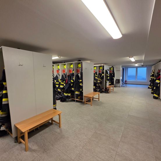 Ein leerer Feuerwehrumkleideraum mit Reihen von Schließfächern, Helmen und Uniformen. Holzbänke und ein Fenster sind ebenfalls vorhanden.