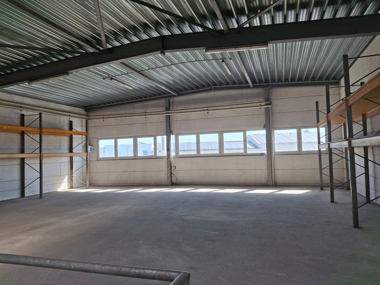Ein leeres Lager mit Metalldach und Glasfenstern. Ein Metallregal steht links.