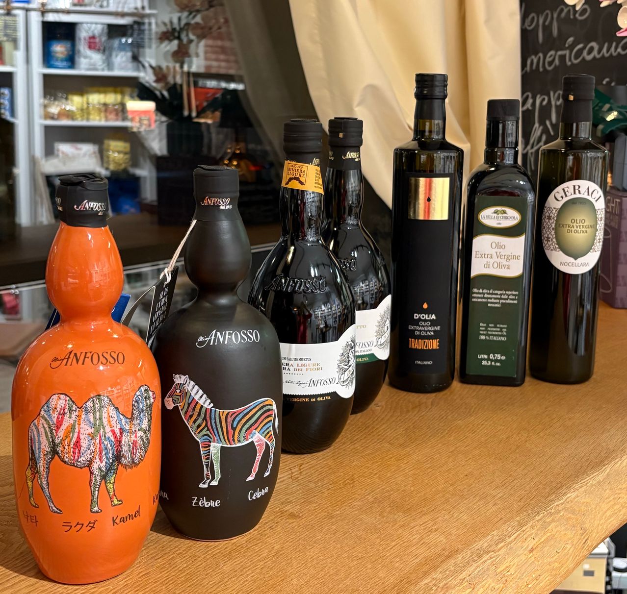 Auf einem Holztisch sind verschiedene Flaschen ausgestellt, darunter zwei mit 'Anfosso' beschriftet, die Kamel- und Zebra-Designs tragen, sowie mehrere Flaschen Olivenöl.