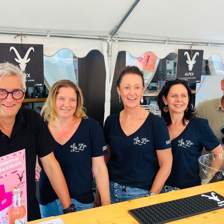 Vier Personen stehen hinter einer Theke in einem Zelt und lächeln für ein Foto. Drei Frauen tragen passende marineblaue Shirts mit weißem Text. Ein Mann in einem schwarzen Shirt hält ein Buch. Dahinter ist ein weißes Zelt mit schwarzen Schildern zu sehen.