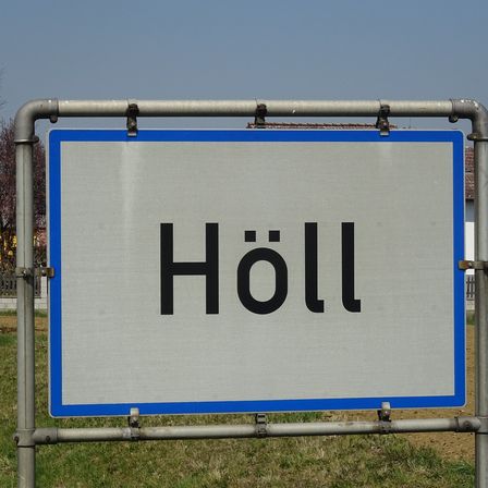 Ein Straßenschild mit dem Wort 'Höll' darauf. Das Schild ist grau mit einem blauen Rand und an einem Metallrahmen befestigt.
