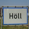 Ein Straßenschild mit dem Wort 'Höll' darauf. Das Schild ist grau mit einem blauen Rand und an einem Metallrahmen befestigt.