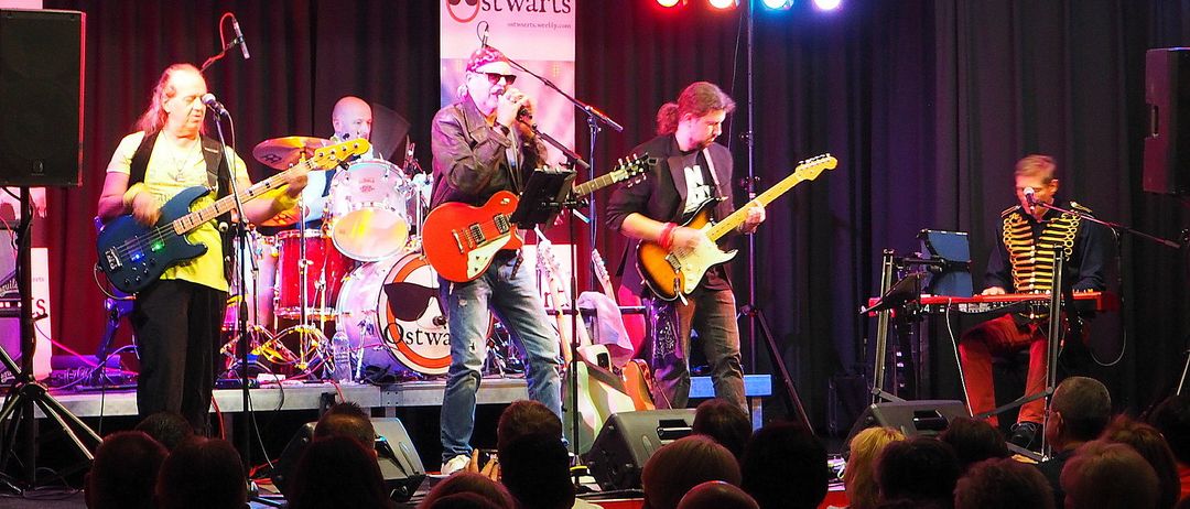 Eine Rockband tritt auf der Bühne auf, mit einem Banner im Hintergrund, das 'stwarts' besagt. Der Leadsänger singt in ein Mikrofon, während zwei Gitarristen neben ihm spielen. Der Schlagzeuger spielt hinter ihnen, und ein Publikum beobachtet von vorne.