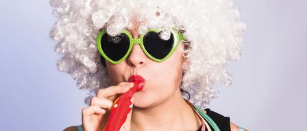 Eine Frau mit weißen, lockigen Haaren, herzförmigen Sonnenbrillen und rotem Lippenstift bläst mit einem roten Blase-Wand Blasen und trägt ein schwarzes Oberteil und bunte Bänder.