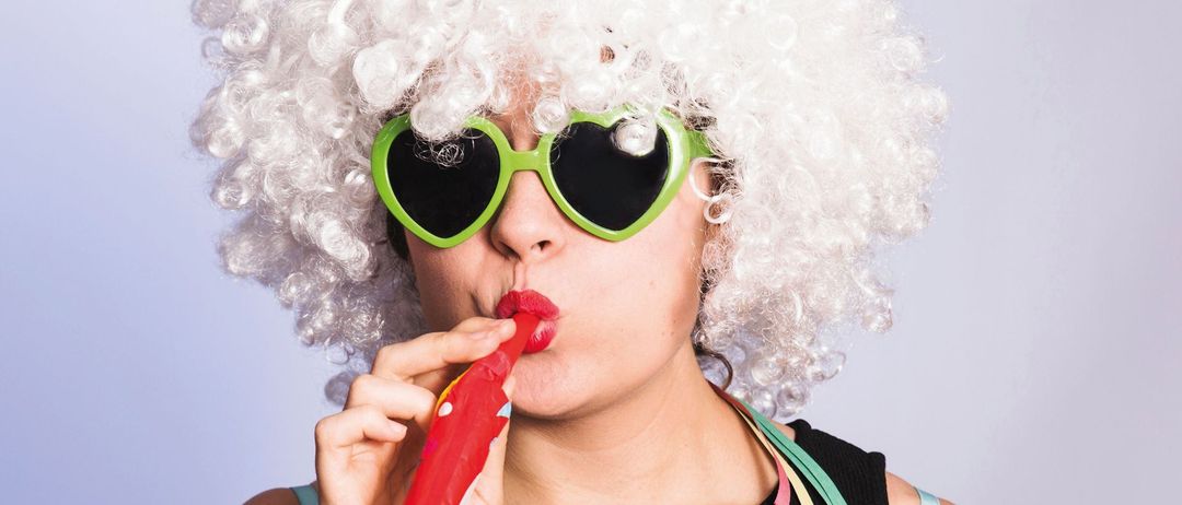 Eine Frau mit weißen, lockigen Haaren, herzförmigen Sonnenbrillen und rotem Lippenstift bläst mit einem roten Blase-Wand Blasen und trägt ein schwarzes Oberteil und bunte Bänder.