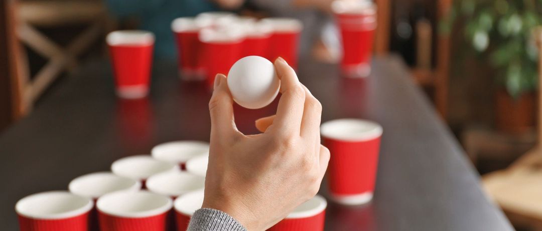 Eine Hand hält einen Tischtennisball über einem Tisch mit vielen roten Bechern, wahrscheinlich in einem Spiel.