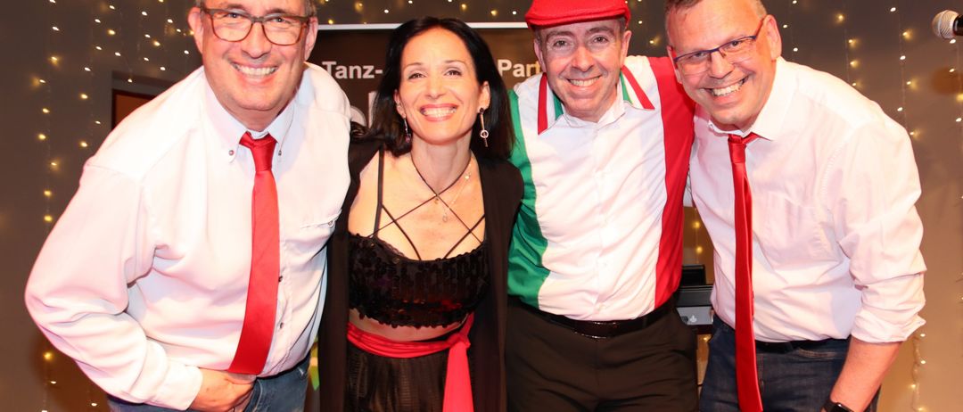 Vier Personen, zwei Männer mit roten Krawatten und einer mit rotem Hut, zusammen mit einer Frau in Schwarz, posieren für ein Foto in einer festlichen Umgebung.