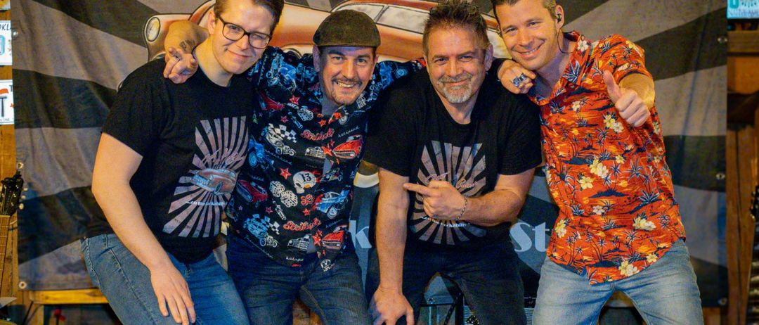 Vier Männer stehen auf einer Bühne mit einem Banner im Hintergrund. Das Banner steht für Gotty & the Cruisers. Die Männer lächeln und posieren für ein Foto.