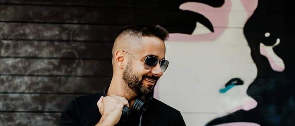 Ein lächelnder Mann in Sonnenbrille und Kopfhörern steht vor einer bunten Graffiti-Wand. Er trägt ein schwarzes T-Shirt mit weißem Grafikdruck.